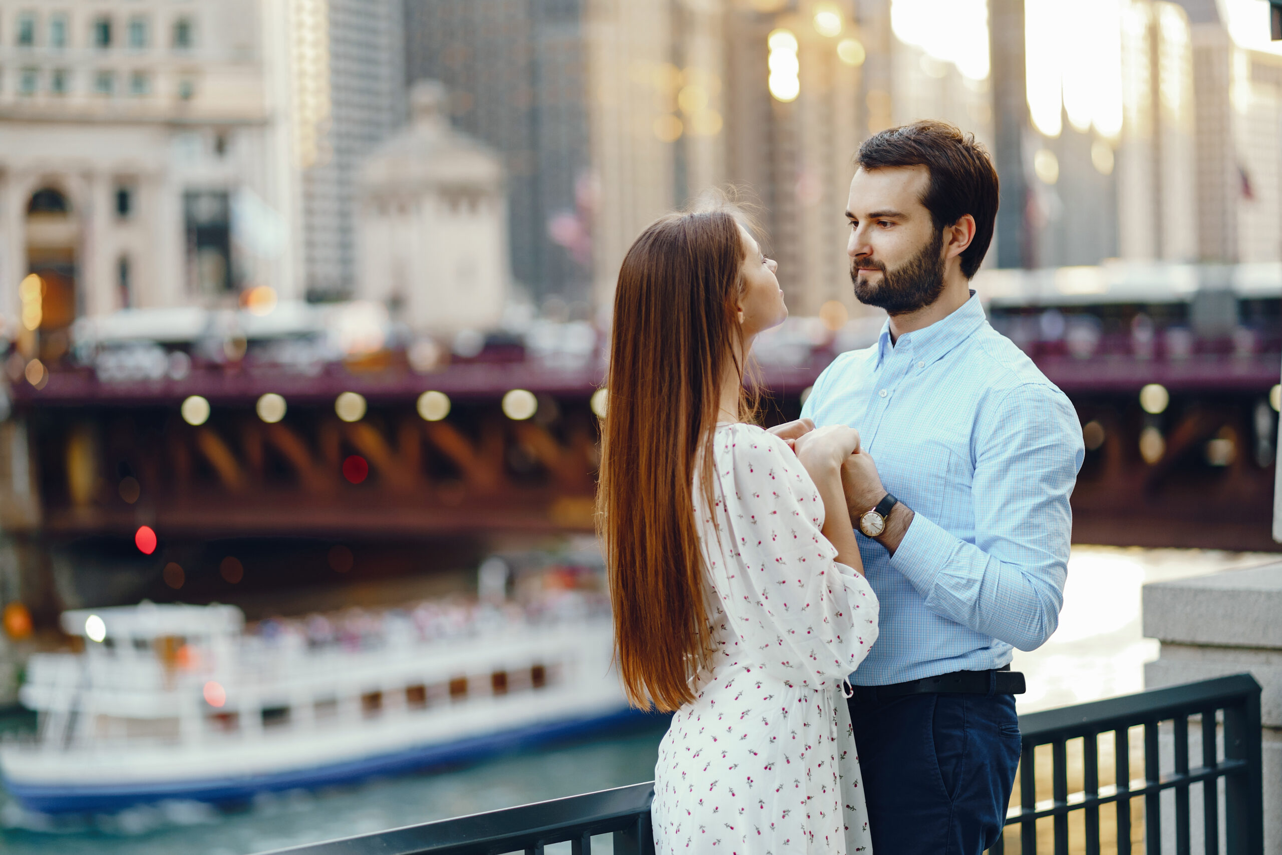 beautiful long-haired girl in summer dress with her handsome husband in white shirt and pants standing in sunny Chicago near river